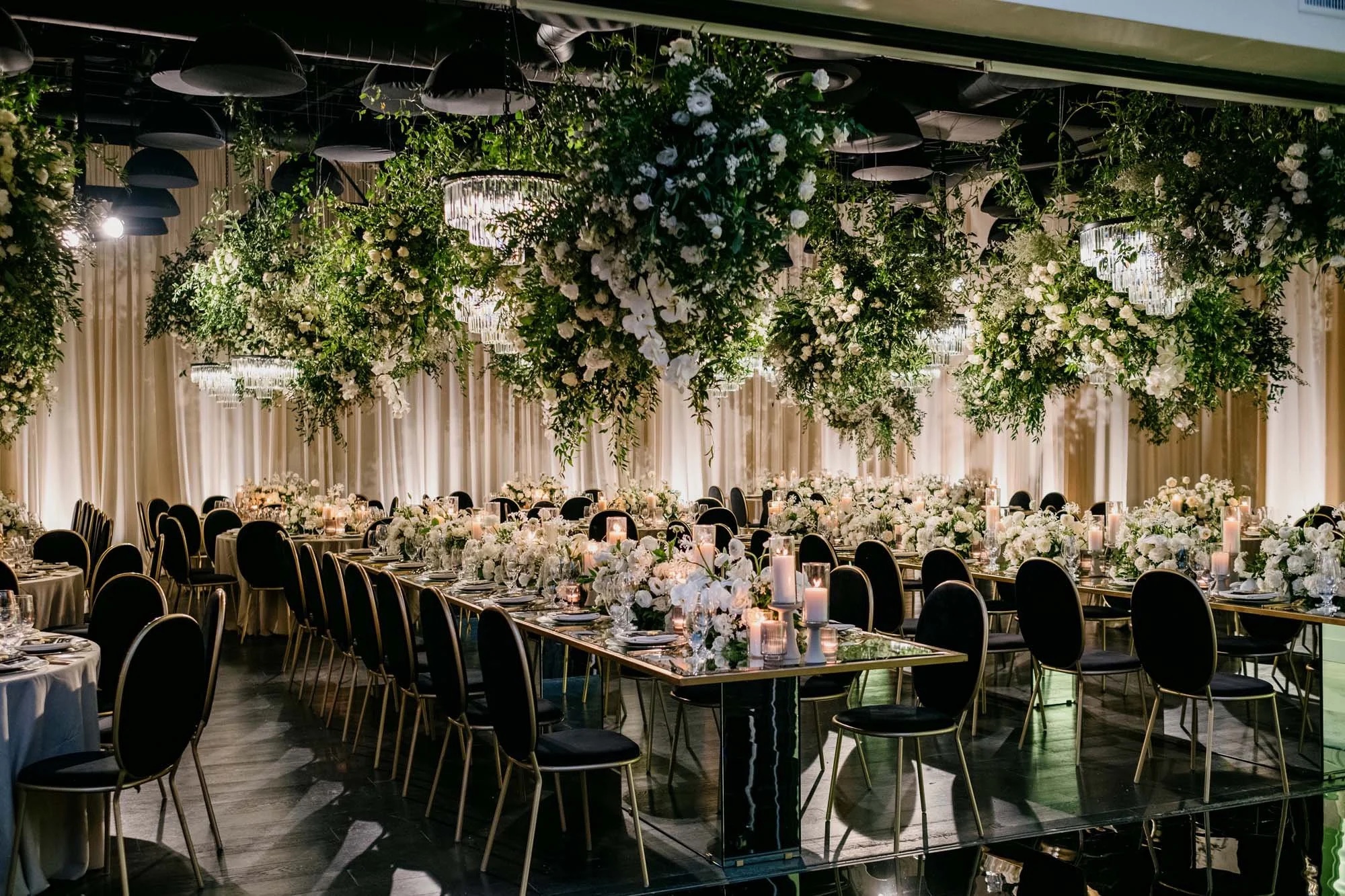Long reception tables beneath hanging greenery and white flowers, with candles and gold edged mirrored tables, About Details Details