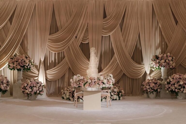Champagne coloured draped backdrop with a tiered cake and pink floral arrangements, About Details Details
