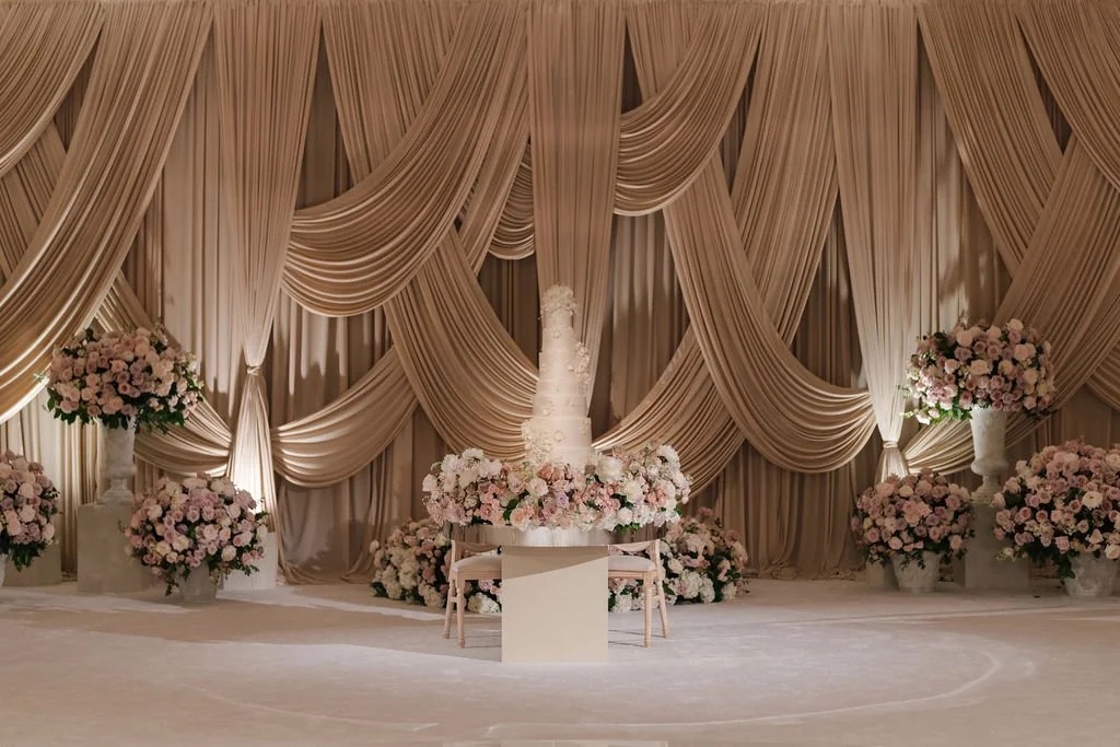 Champagne coloured draped backdrop with a tiered cake and pink floral arrangements, About Details Details