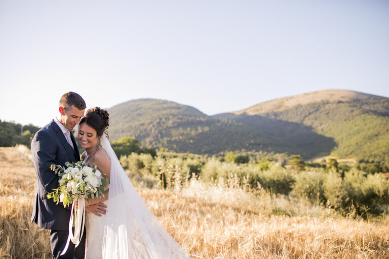 Couple embracing in dry golden fields with wooded hills behind, Alessandro Mari Photography