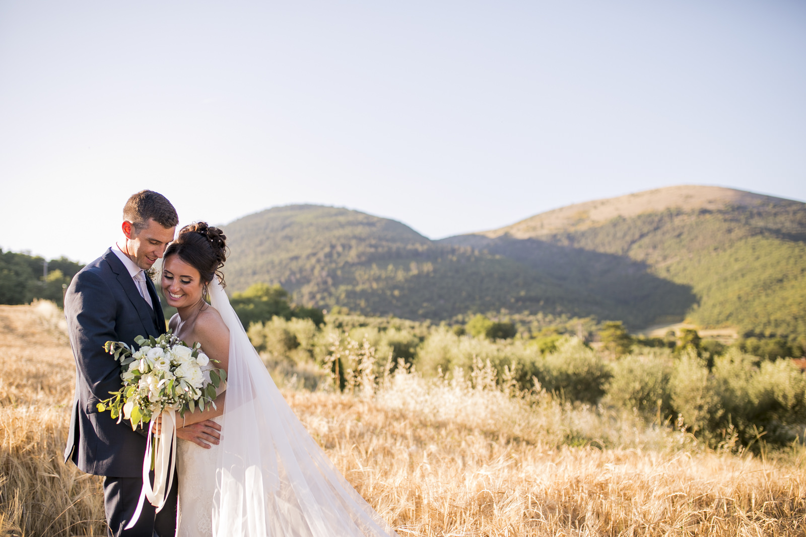 Couple embracing in dry golden fields with wooded hills behind, Alessandro Mari Photography