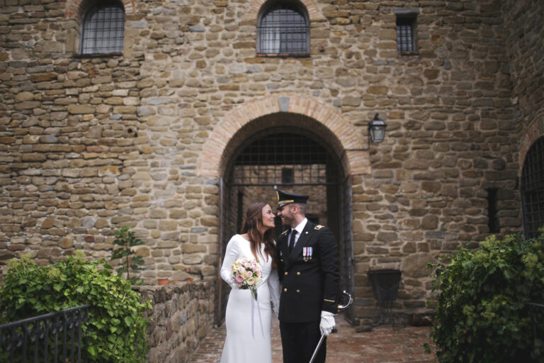 Bride and groom in uniform at a medieval stone gateway, Alessandro Mari Photography