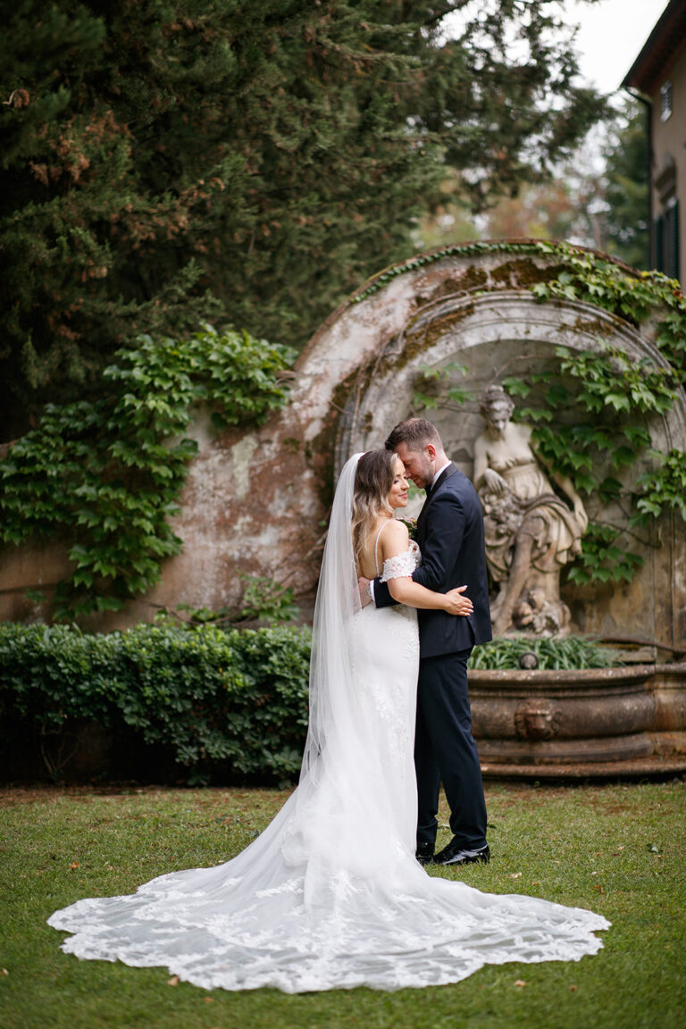 Couple in a garden with a long lace veil by a stone fountain, Alessandro Mari Photography