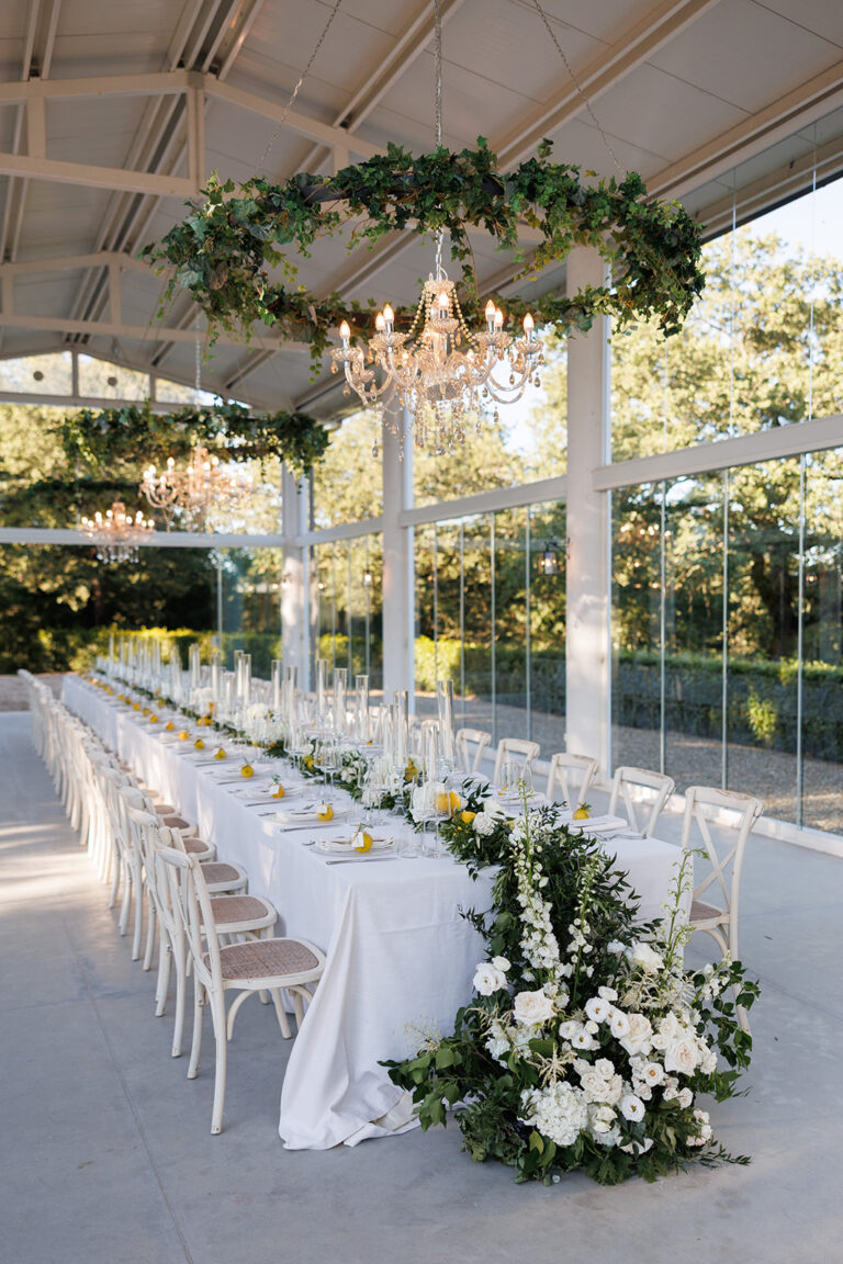 Long reception table under a glass-walled pavilion with a greenery chandelier, Alessandro Mari Photography
