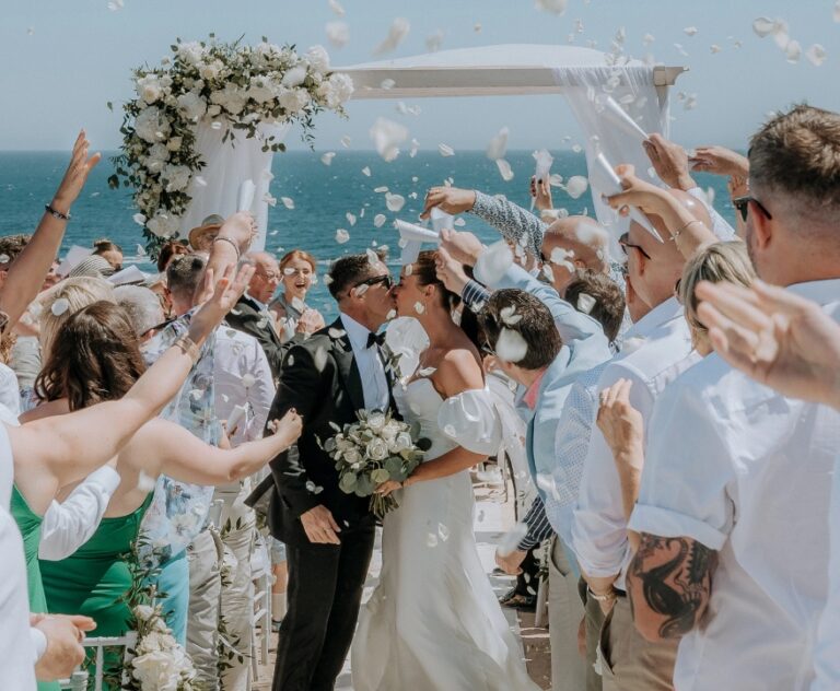 Guests throwing petals as the couple leave the ceremony under a floral arch by the sea, Algarve Wedding Planners