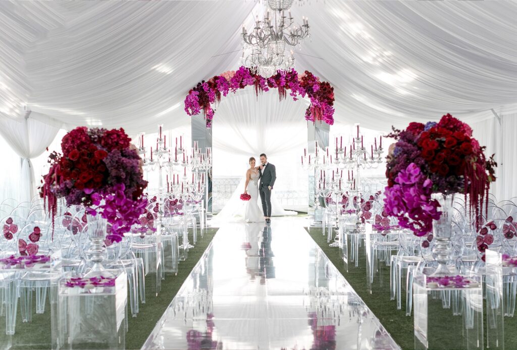A ceremony aisle with a magenta floral arch and clear candelabra in a white draped space