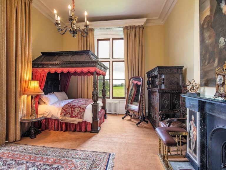 A bedroom with a canopied four-poster in cream and gold, framed by a window to the grounds, Allerton Castle