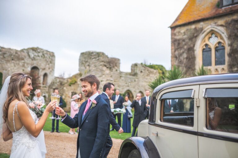 A couple toasting with Champagne beside a vintage car, guests and the old chapel behind, Amberley Castle