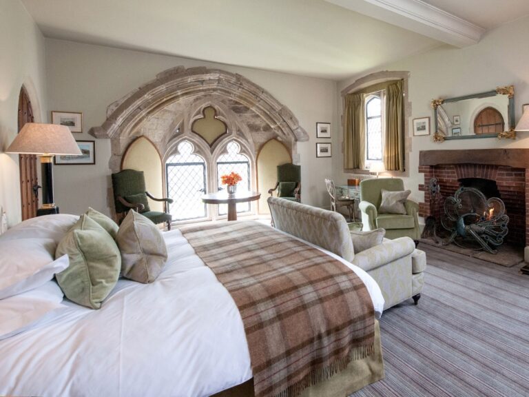 A castle bedroom with a stone-arched window, a fireplace and armchairs, Amberley Castle