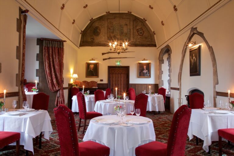 A vaulted stone dining hall with a painted armorial panel, a chandelier and candlelit tables set for dinner, Amberley Castle