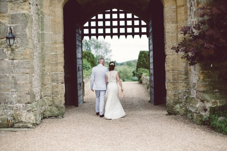 A bride and groom walking hand in hand beneath the portcullis, out toward the gardens, Amberley Castle