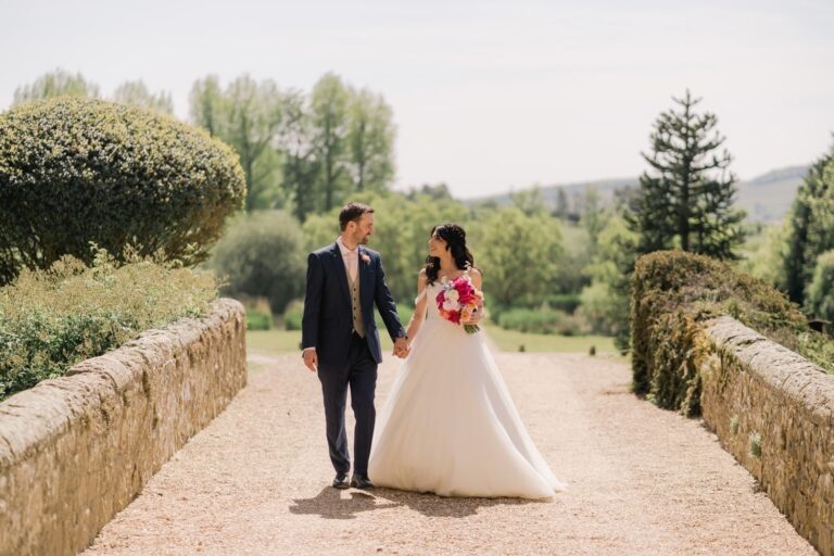 A bride and groom on a stone causeway across the moat, the hills rising behind, Amberley Castle