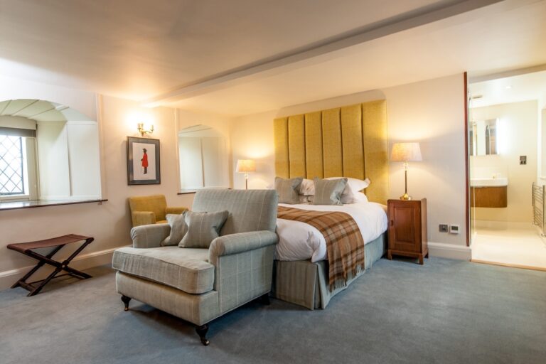 A guest room with a large bed dressed in a tartan throw and a fireside armchair, Amberley Castle
