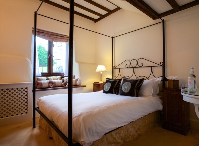 A four-poster bedroom under a beamed ceiling, with a cushioned window seat, Amberley Castle