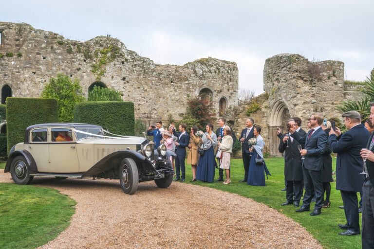 A vintage cream car on the gravel drive as guests applaud the couple, Amberley Castle