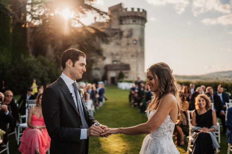 The couple holding hands at the ceremony, the castle keep and a low sun behind, Anita Galafate