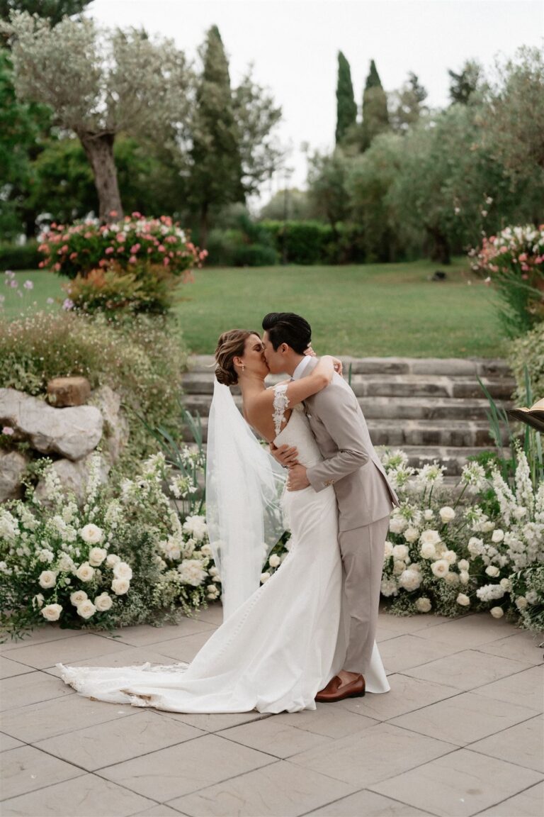 The couple kissing in an olive garden, white florals banked at the foot of the steps, Anita Galafate