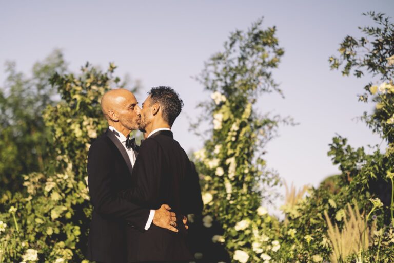 Two grooms kissing in a garden, white roses and greenery banked around them, Anita Galafate