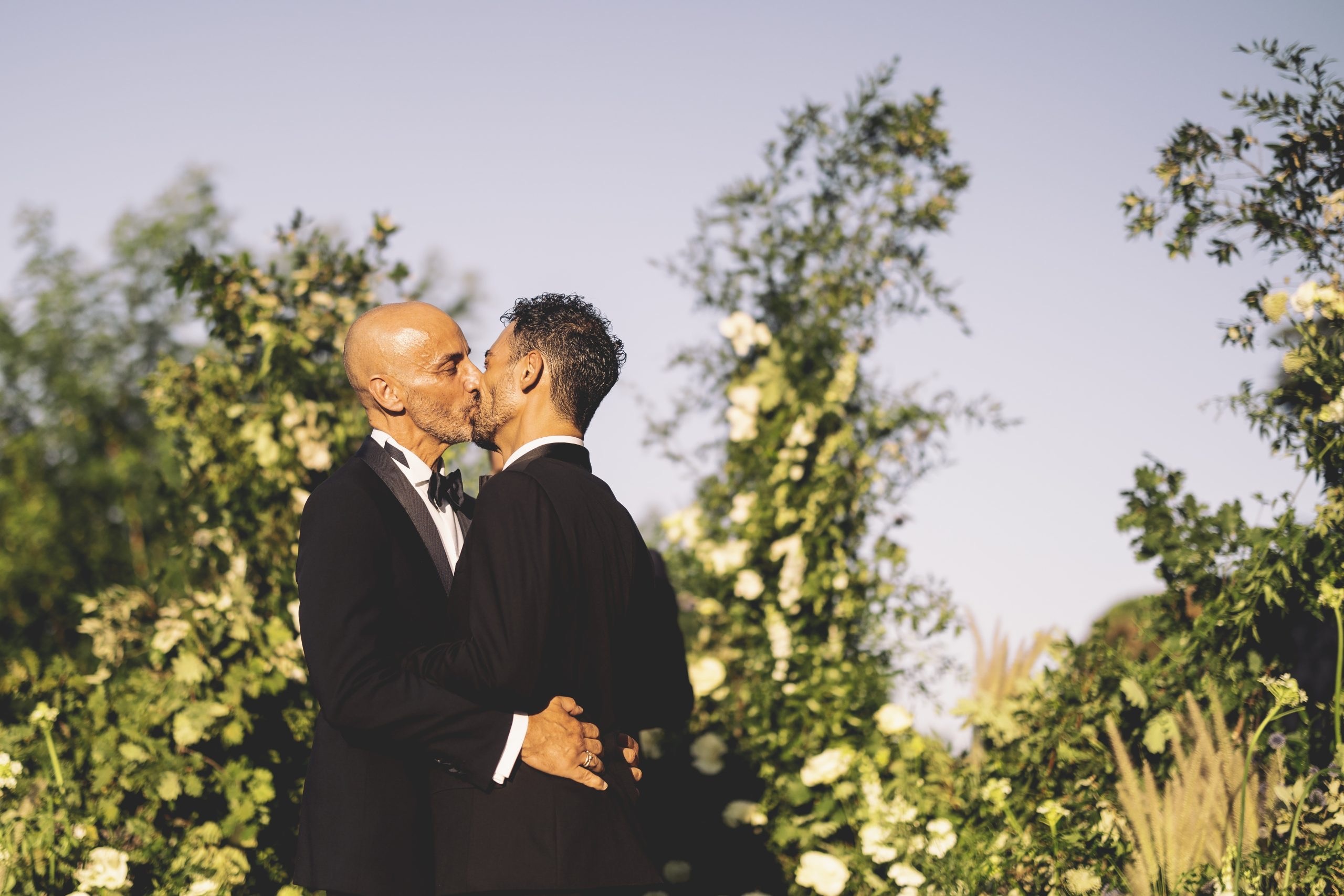 Two grooms kissing in a garden, white roses and greenery banked around them, Anita Galafate