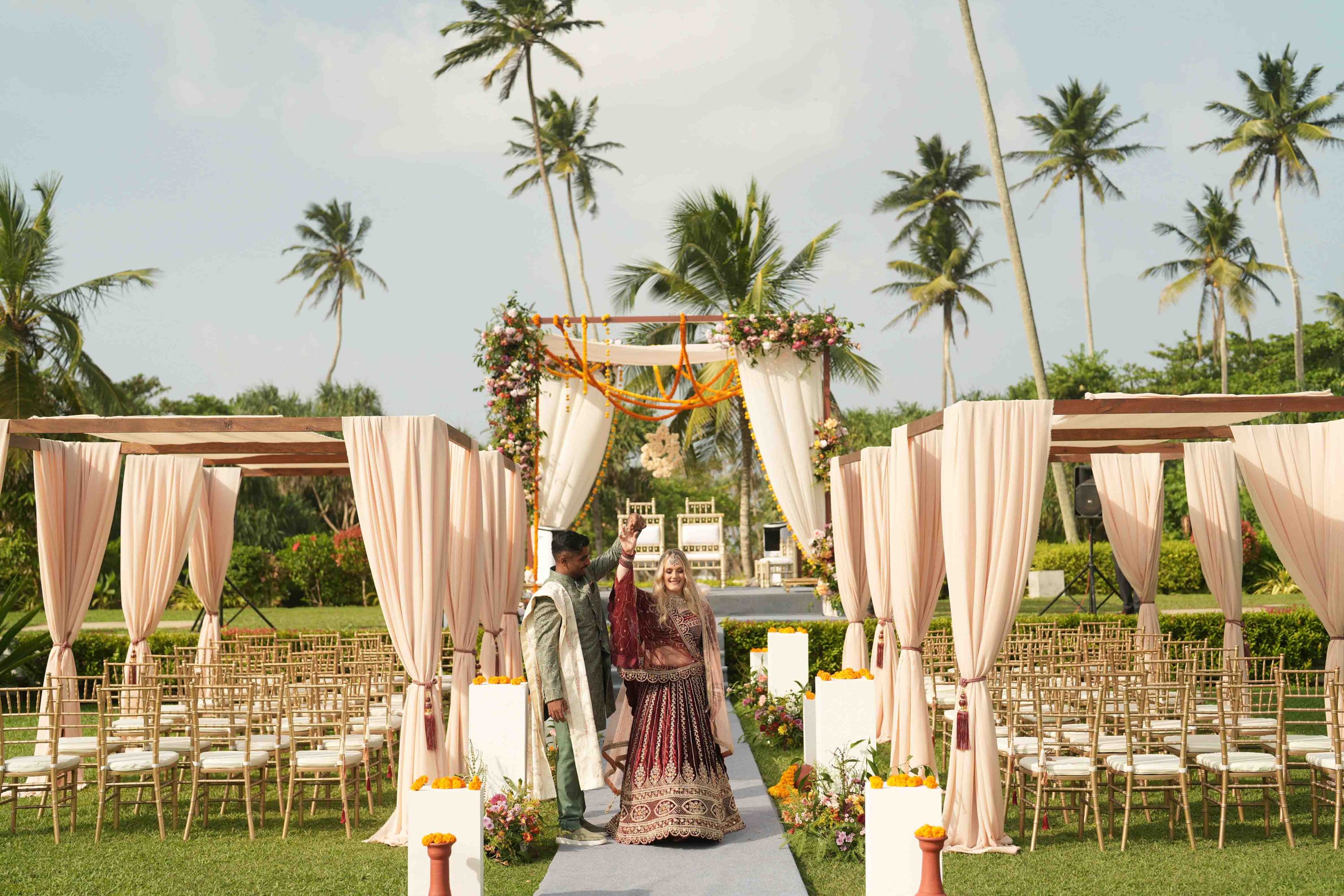 A draped mandap and rows of gold chairs on a lawn among palm trees, with the bride walking up the aisle, Anywhera Events The Destination Wedding Planner in Sri Lanka