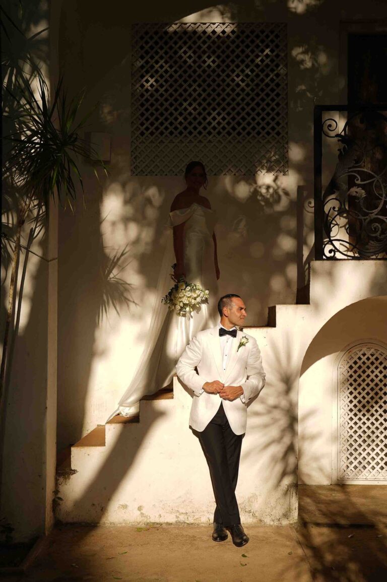A groom in a white dinner jacket leaning against a sunlit stucco staircase with the bride behind, Anywhera Events The Destination Wedding Planner in Sri Lanka