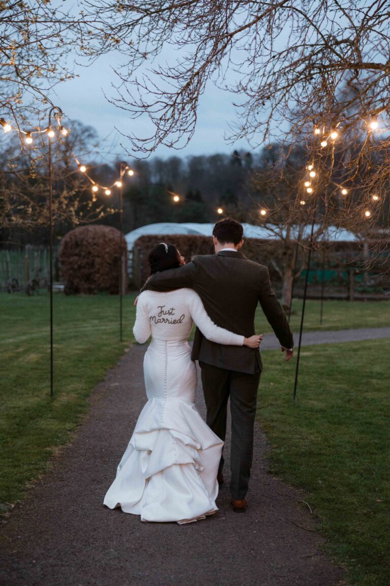 Newly-weds walking a festoon-lit path through the grounds at dusk, Askham Hall
