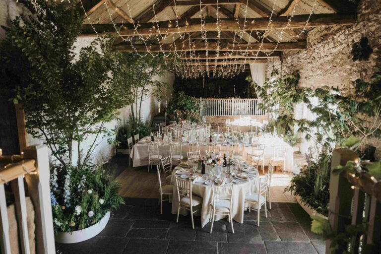 Bank Barn set for the wedding breakfast, with round tables, potted trees and fairy lights across the beams, Askham Hall