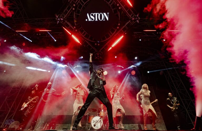 The full band on stage under red lighting with the Aston sign overhead, Aston Band