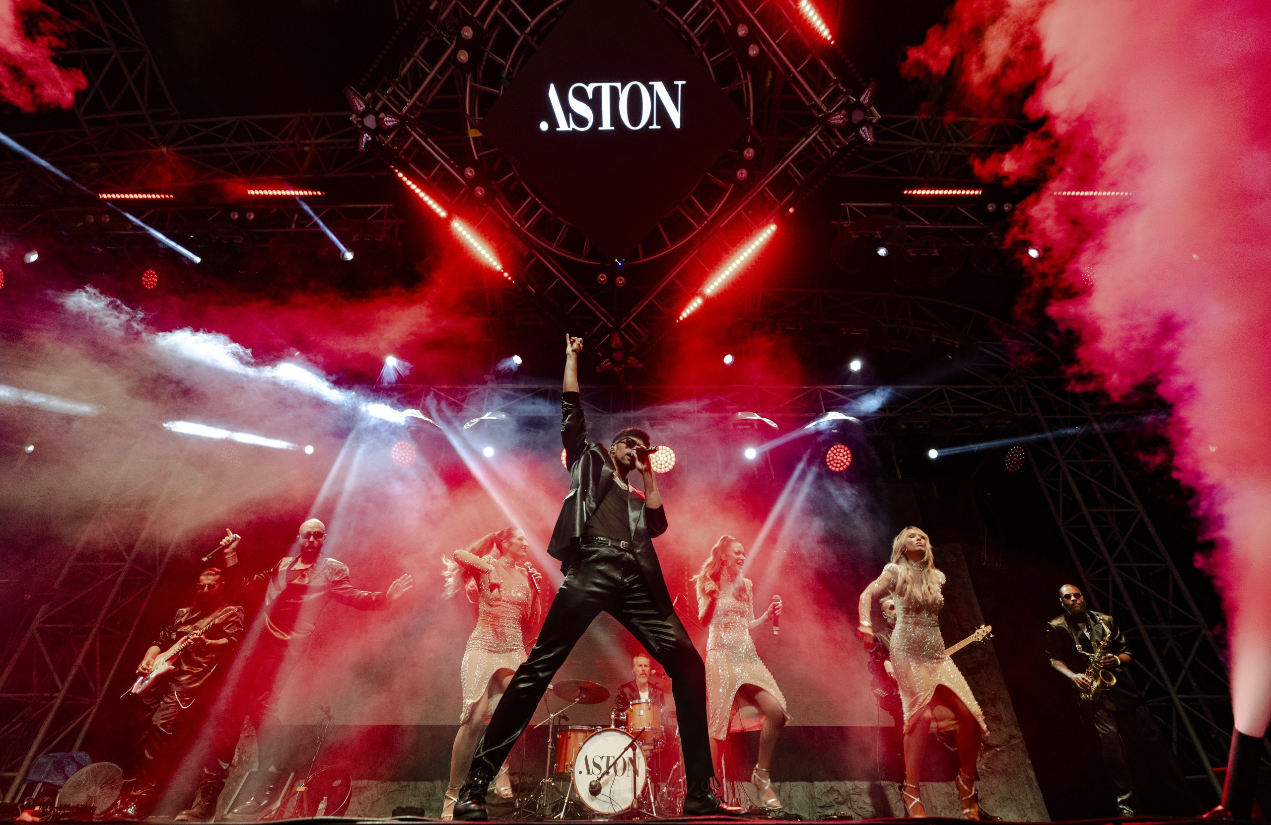 The full band on stage under red lighting with the Aston sign overhead, Aston Band