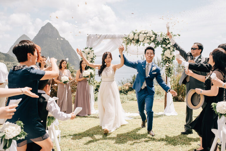 Couple walking out beneath a white floral arch as guests throw confetti, with the Pitons behind, Awesome Caribbean Weddings