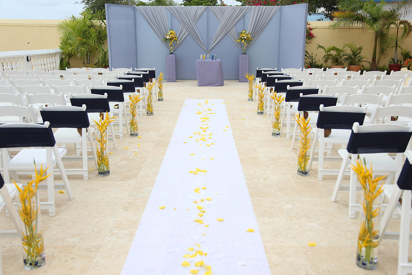 Ceremony set up on a white aisle runner between rows of white chairs, with yellow flowers and a draped backdrop, Awesome Caribbean Weddings