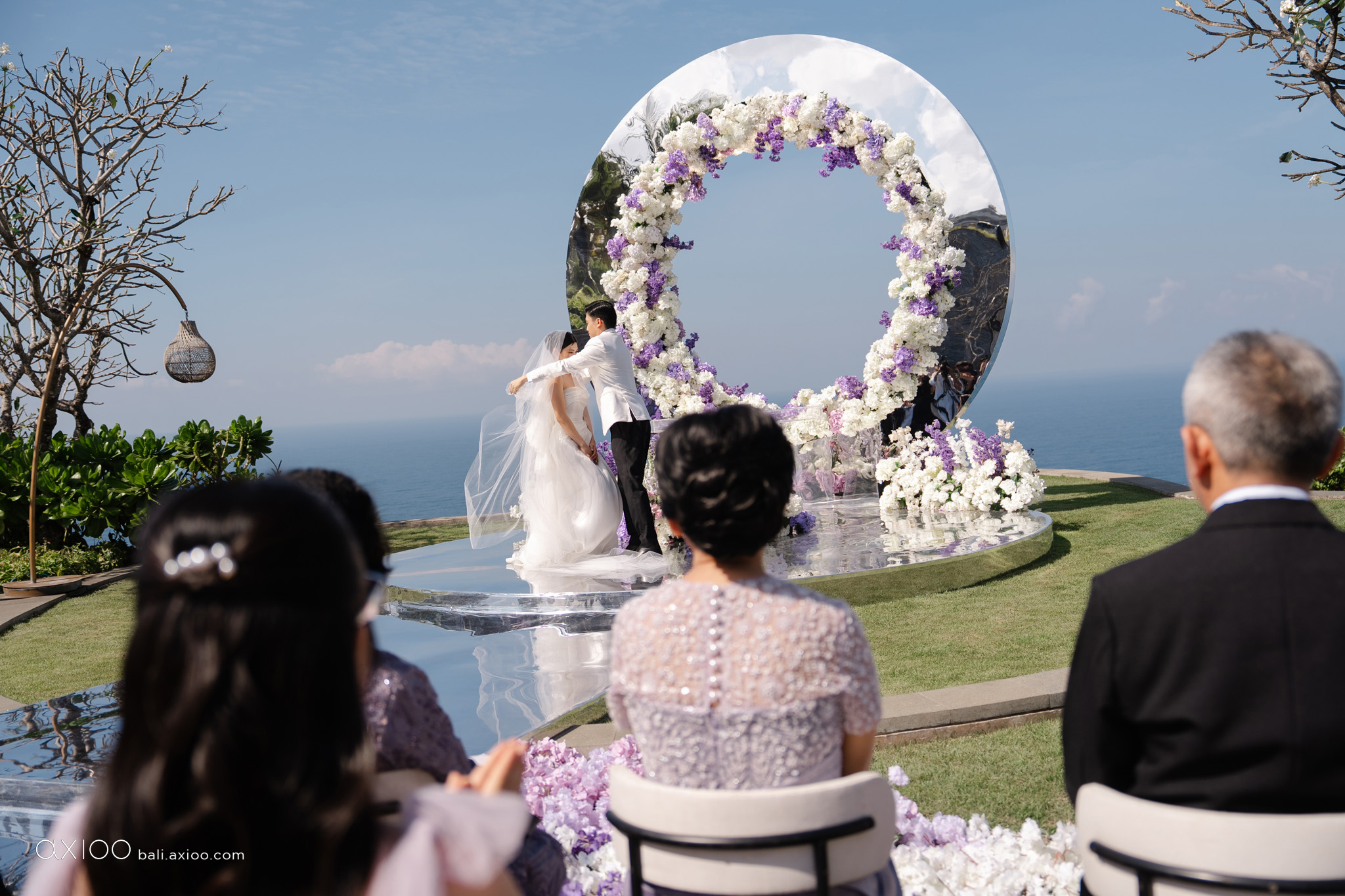 Ceremony beside a circular floral arch with parents watching, ocean beyond, Bali, Axioo Bali