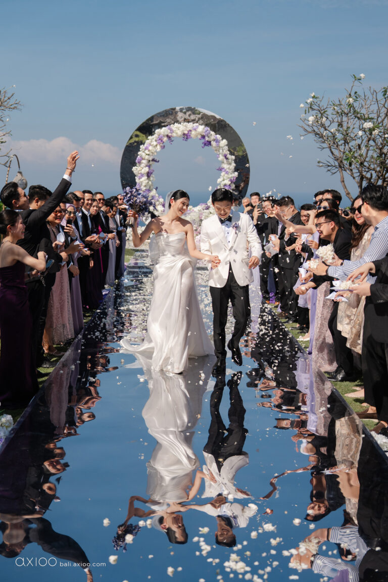 Newlyweds walking a mirrored aisle past guests towards a floral arch, Bali, Axioo Bali