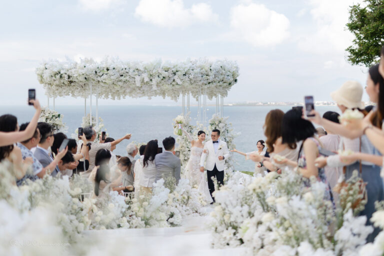 Ceremony beneath a suspended floral canopy with guests filming, ocean beyond, Bali, Axioo Bali