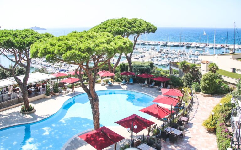 The panoramic pool set among umbrella pines above the marina, burgundy loungers along the water, Baglioni Resort Cala del Porto
