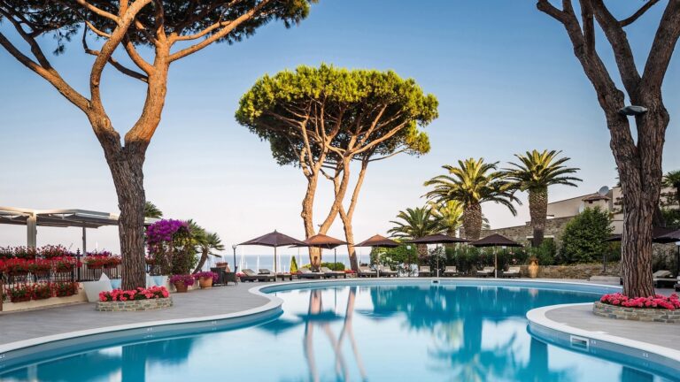 The pool by day, framed by pines and flowering shrubs, the loungers empty in the Tuscan sun, Baglioni Resort Cala del Porto