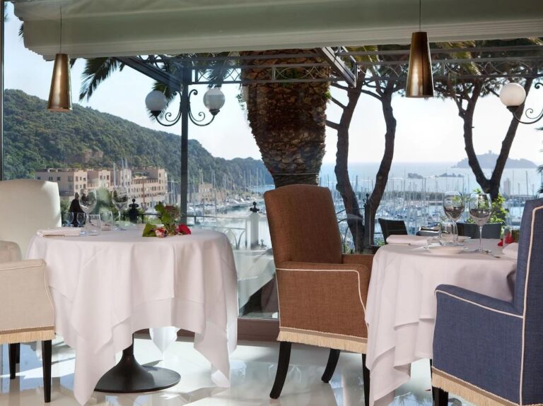 The Belvedere terrace laid with white linen, the marina and pines through the glass, Baglioni Resort Cala del Porto