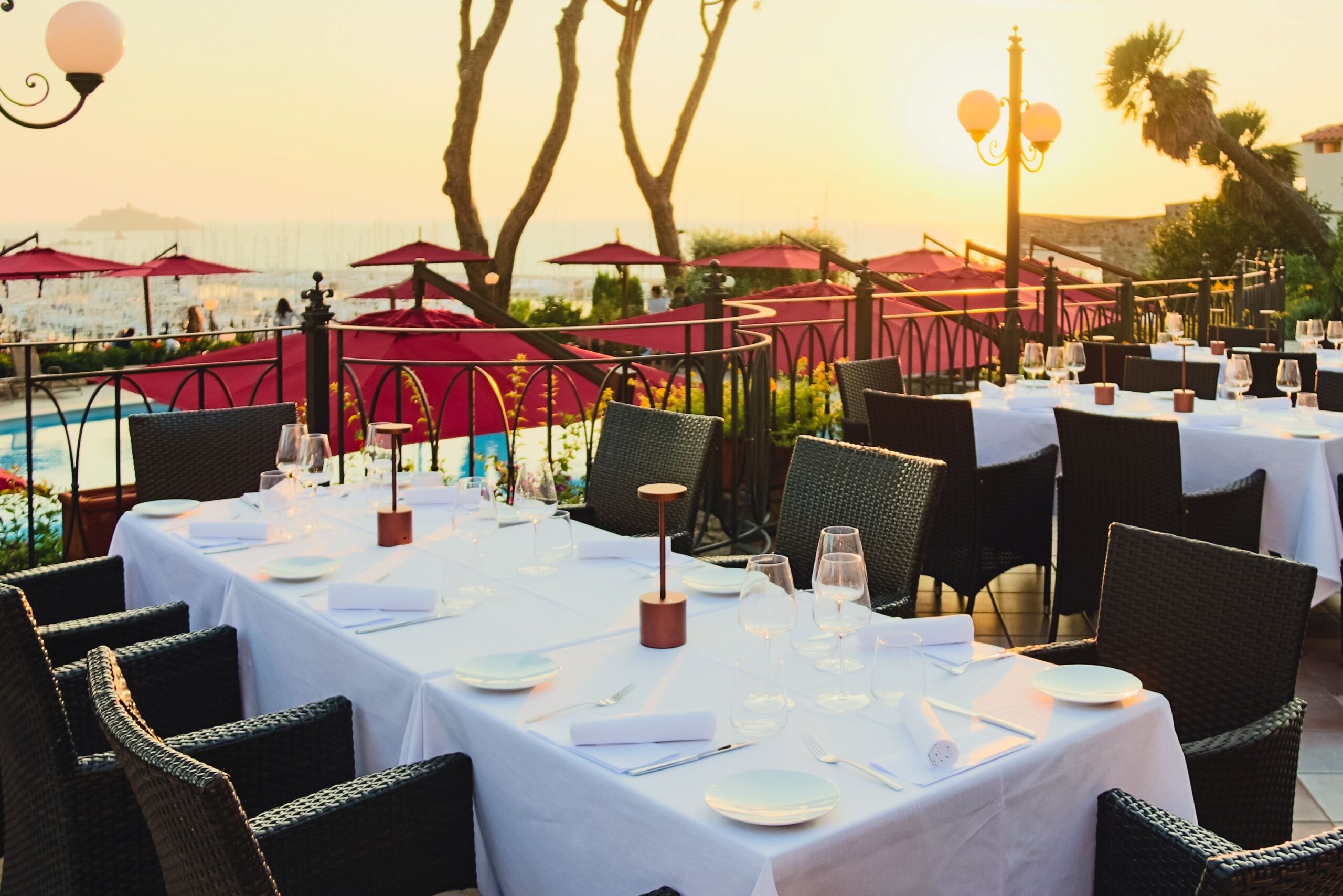 The Sunset Gourmet Terrace laid for dinner in cream linen and burgundy parasols, the marina and sea at golden hour, Baglioni Resort Cala del Porto