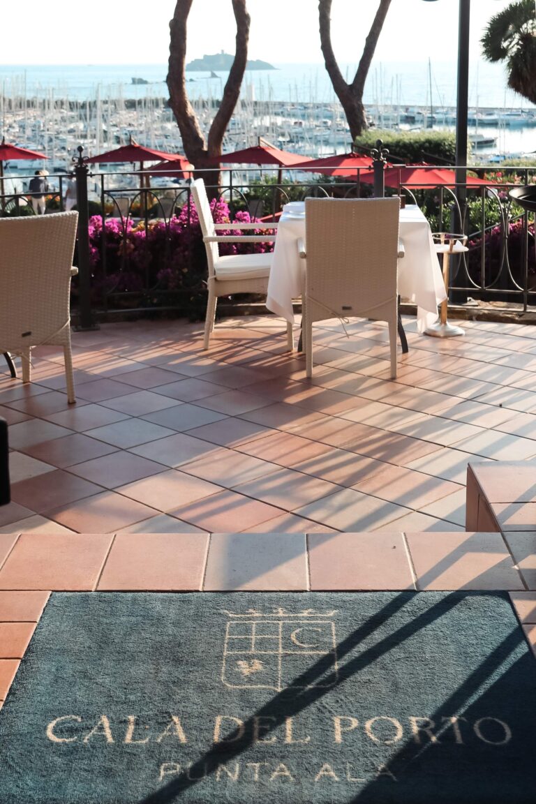 The terrace restaurant set for dinner beneath burgundy parasols, the Cala del Porto crest underfoot, Baglioni Resort Cala del Porto