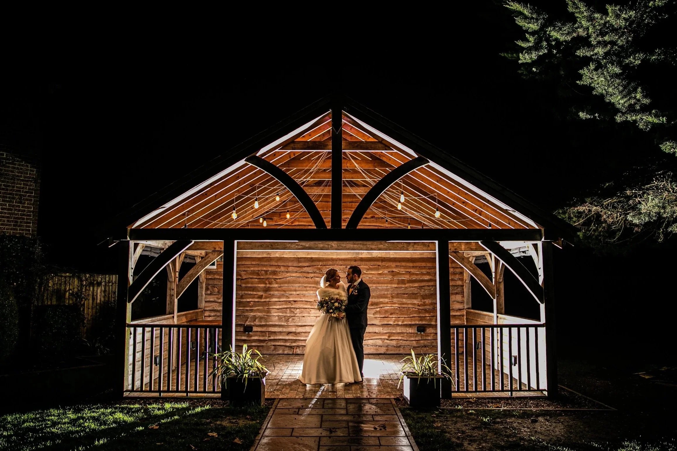 The timber Sylvan Pavilion lit with festoon bulbs at night, a bride and groom standing inside, Balmer Lawn Hotel & Spa