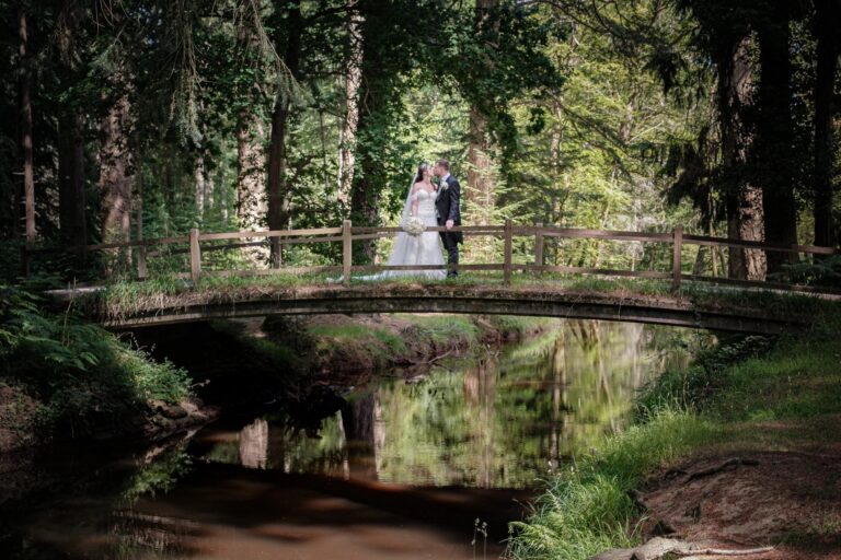 A bride and groom on a wooden footbridge over a stream among tall New Forest trees, Balmer Lawn Hotel & Spa