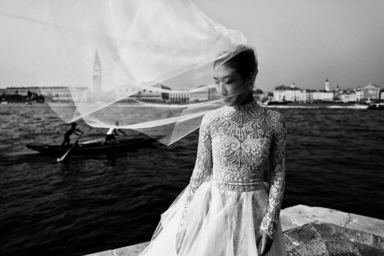 A bride's veil lifting in the wind on the Venetian lagoon, a gondola passing behind, Barbara Zanon Photography