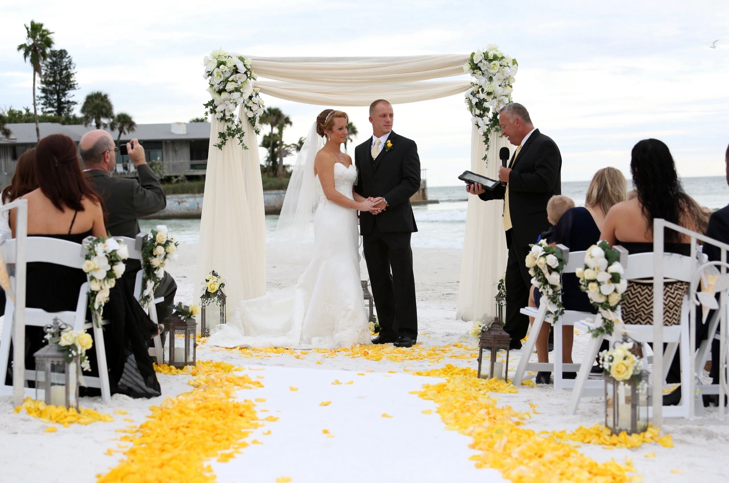 Ceremony beneath a draped white arch with a yellow petal aisle, on a Sarasota beach, Beach Breeze Weddings