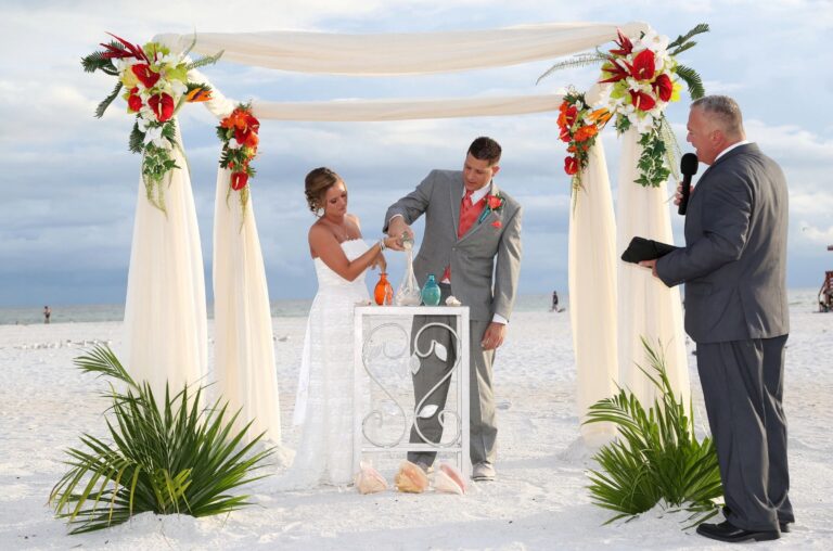 Sand ceremony under an arch of red, orange and white flowers on the beach, Beach Breeze Weddings
