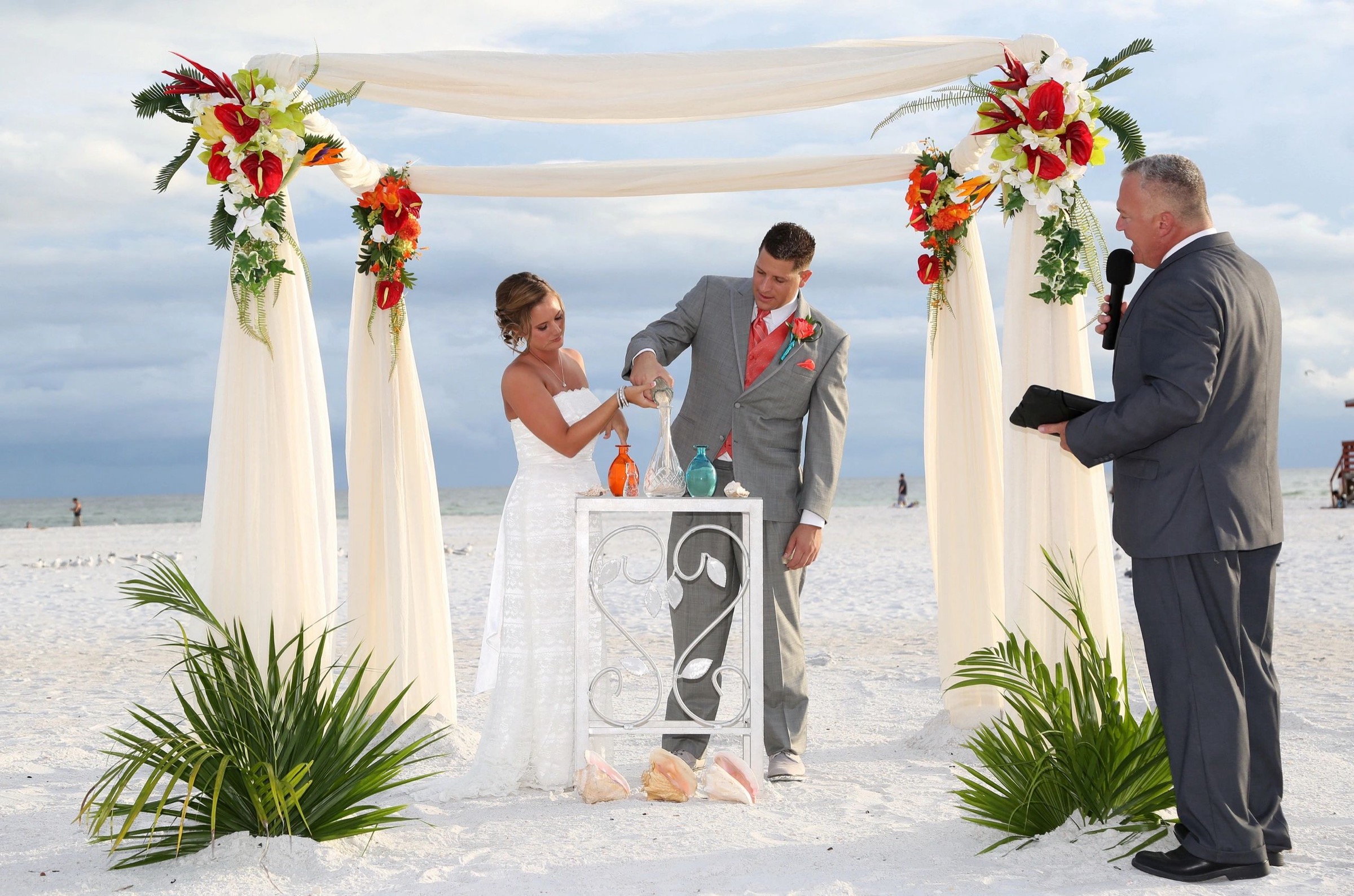 Sand ceremony under an arch of red, orange and white flowers on the beach, Beach Breeze Weddings