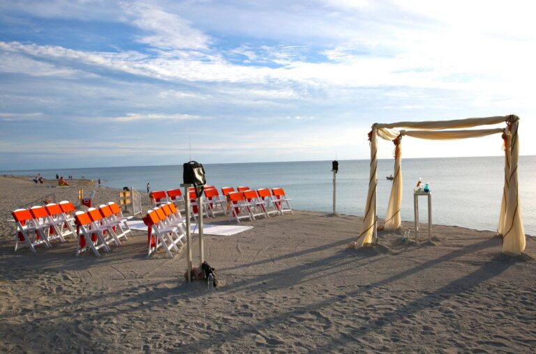 Orange chairs facing a draped arch on the sand at the water's edge, Beach Breeze Weddings