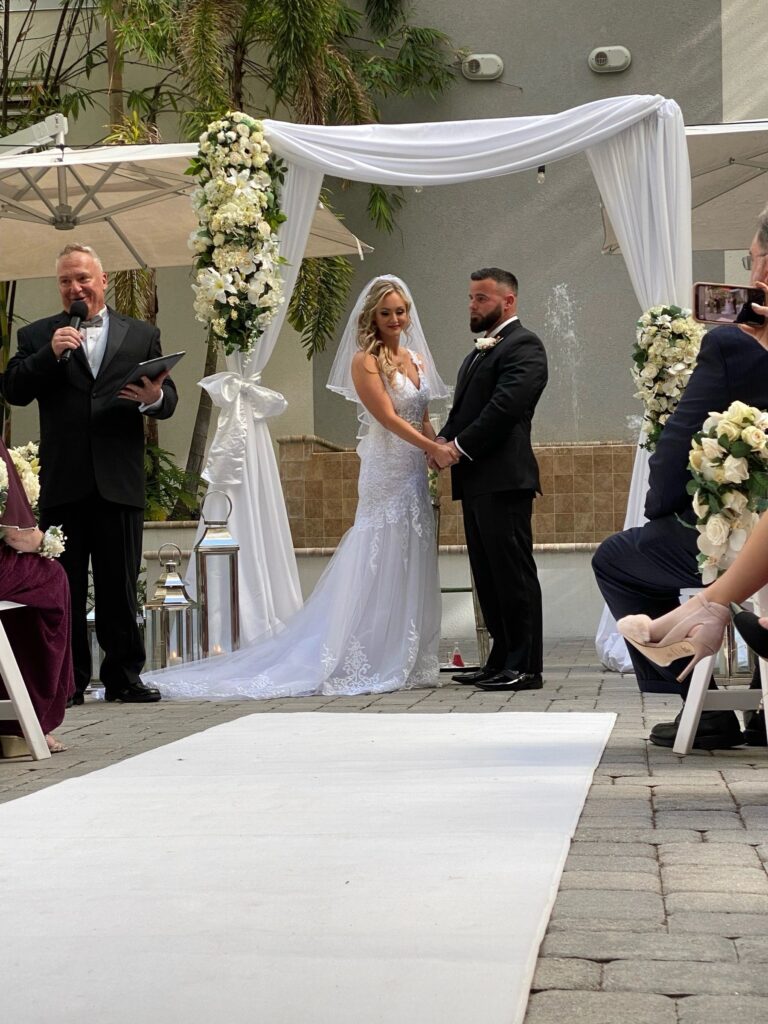 Courtyard ceremony with a white aisle runner and a flower-dressed arch, Beach Breeze Weddings