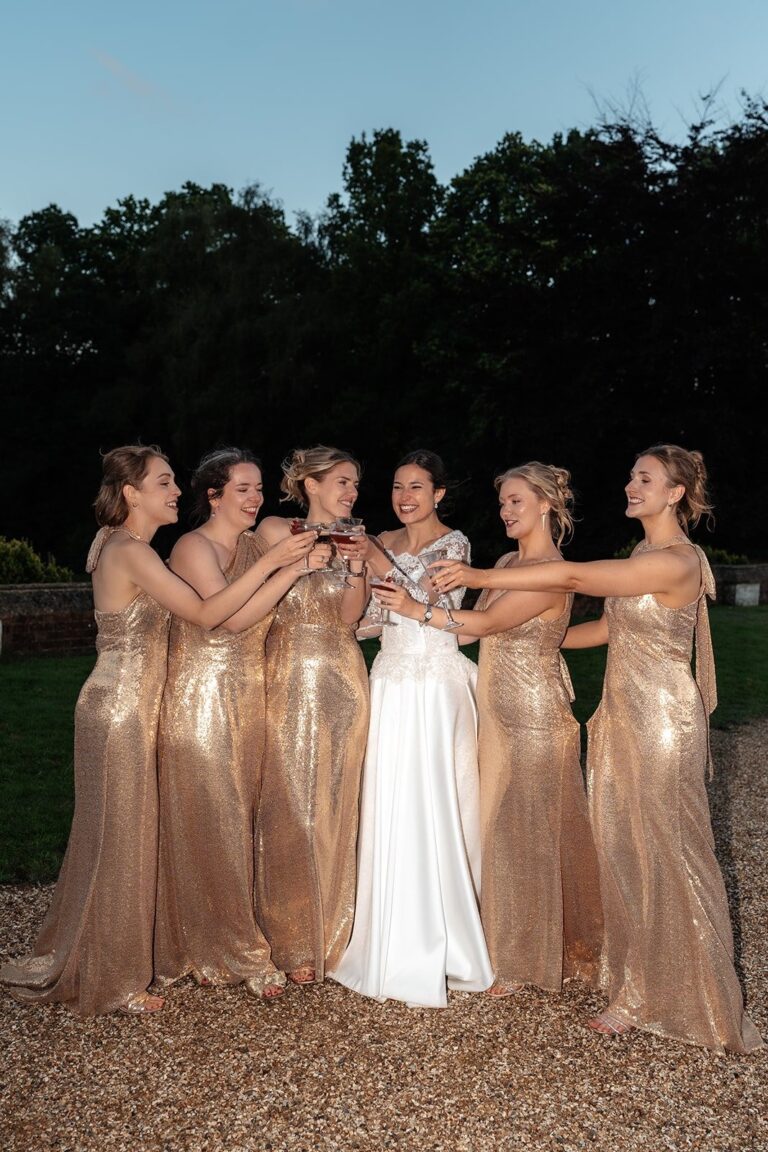 A bride in a white gown raising a glass with five bridesmaids in bespoke gold sequin dresses outdoors, Beatrice Espinosa Bridal