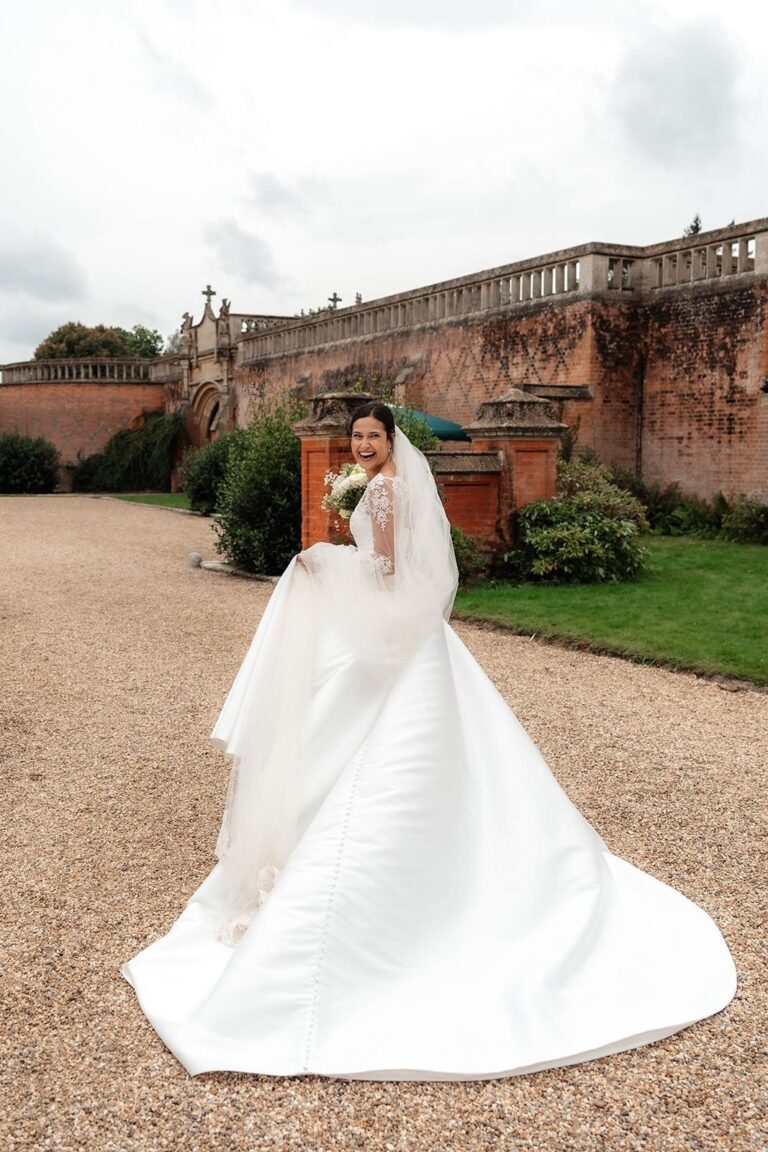 A bride in a satin gown with a long train and veil on a gravel drive beside a manor wall, Beatrice Espinosa Bridal