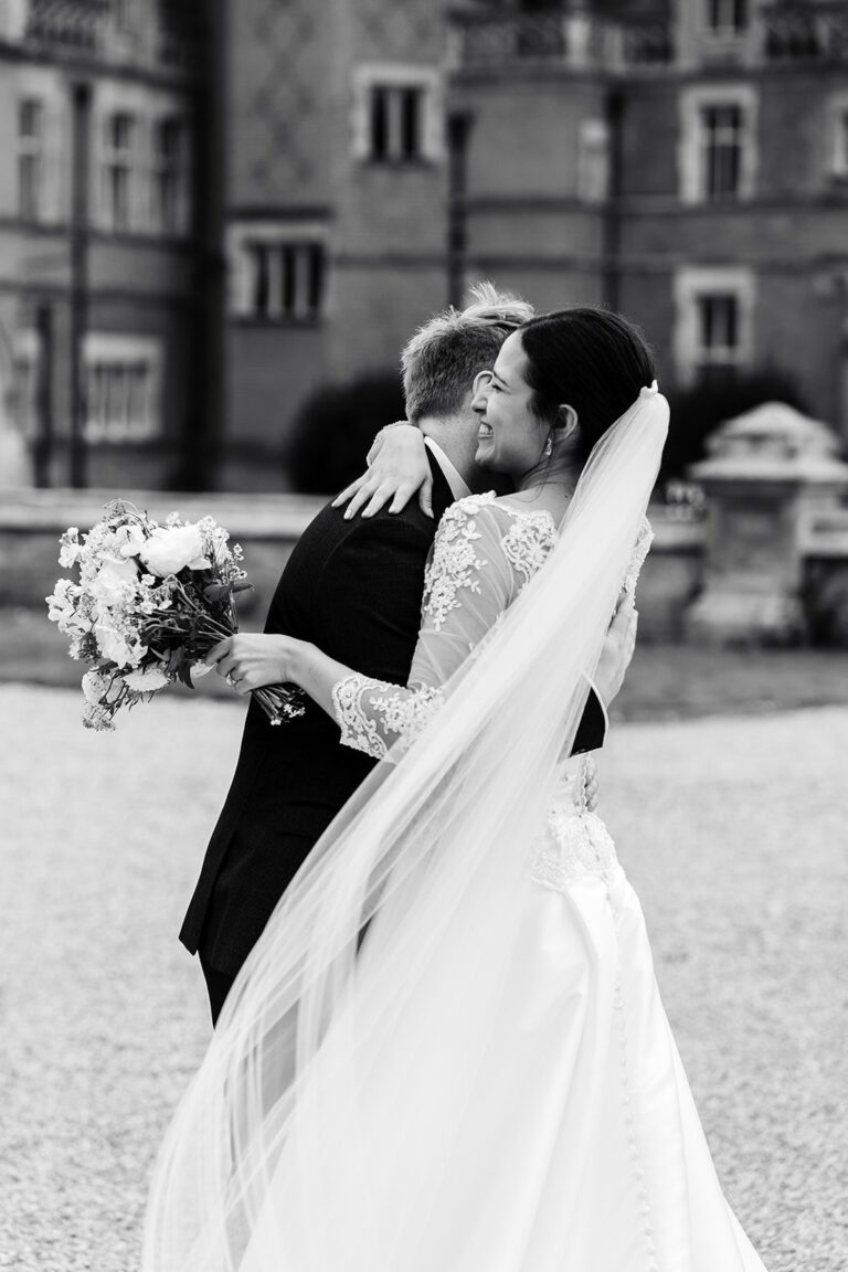 A black and white portrait of a couple embracing, the bride in a lace-sleeved gown with a long veil, Beatrice Espinosa Bridal